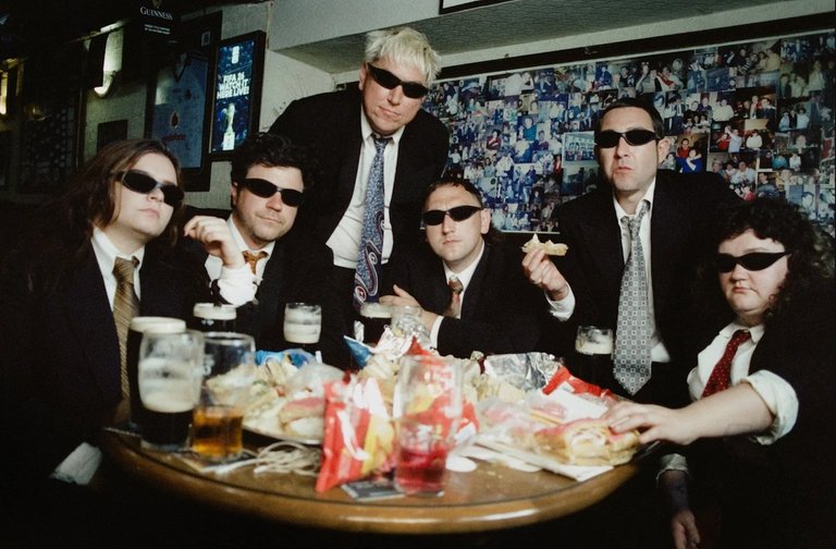 Irish folk band The Mary Wallopers members wearing suits and sunglasses while sitting around a pub table with pints of stout and food
